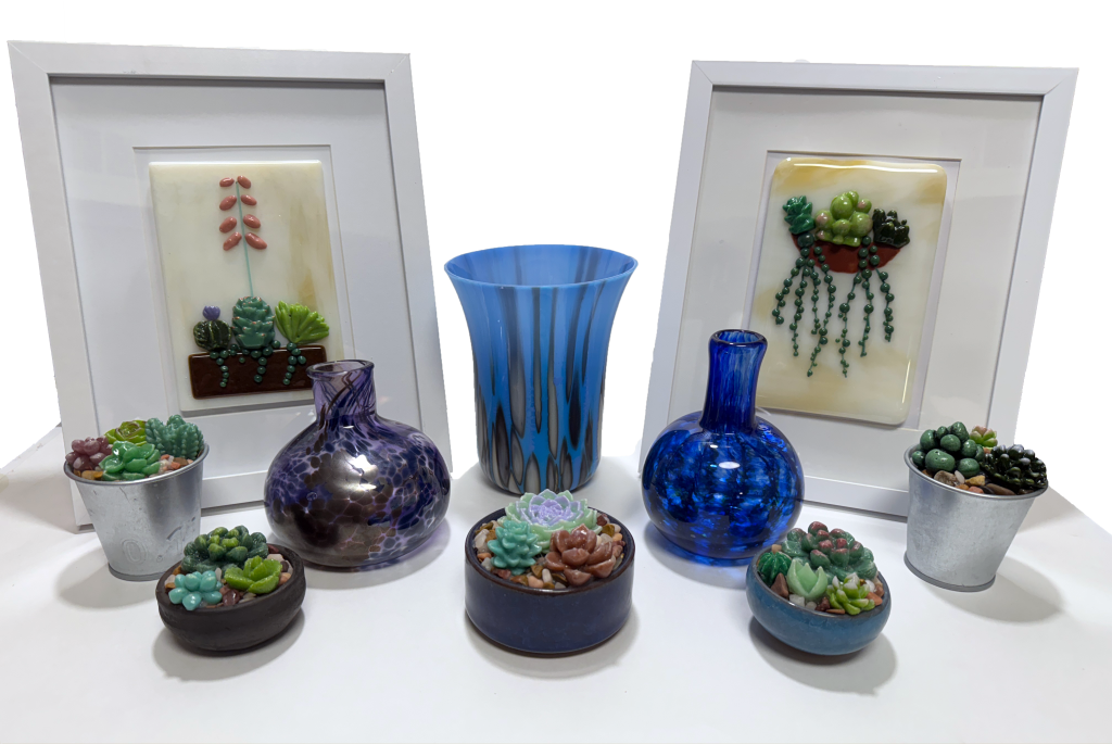 Display for fused and blow glass pieces including a blue and French vanilla streaky vase in the center, two framed wall pieces showing succulents, five succulent gardens in a variety of ceramic or metal pots, and two pulled-neck blown vases, one dark purple with silver speckles and one dark blue with darker blue accents in a ridge pattern.